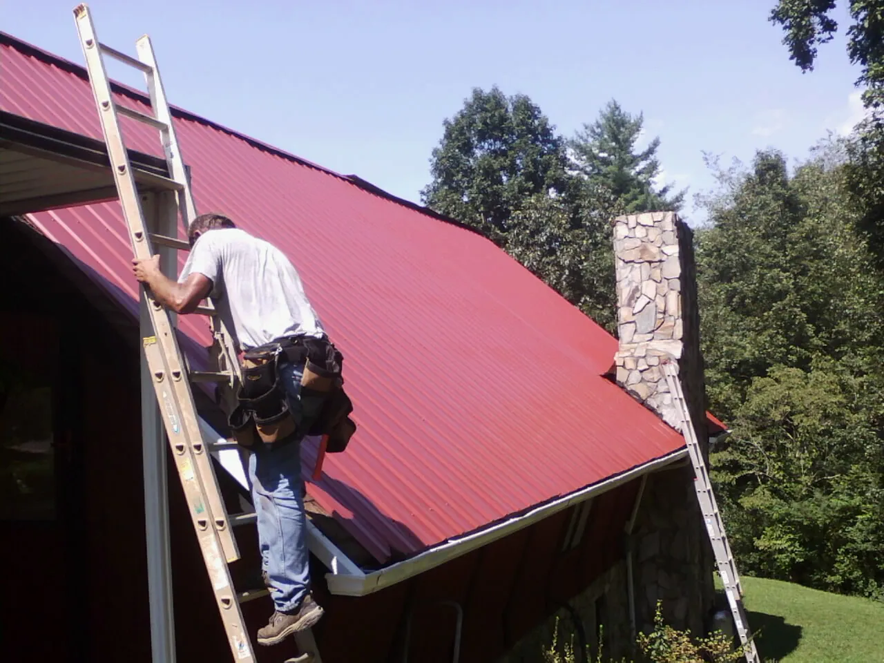 Roofer on a ladder beside a red metal roof and stone chimney — Soffit & Fascia Repair by Roofing Brothers in Parsippany